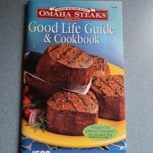 May include: A blue and white cookbook titled "Good Life Guide & Cookbook" by Omaha Steaks. The cover features a close-up of grilled steaks on a yellow plate with vegetables and rosemary.
