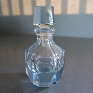 May include: A clear, faceted glass perfume bottle with a matching stopper. The bottle has a geometric design and is filled with a clear liquid. The bottle is approximately 10 cm tall.
