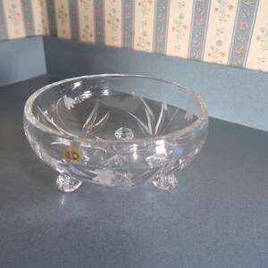 May include: A clear, crystal glass bowl with a floral design etched into the sides. The bowl has three small, rounded feet and a small, round knob in the center. The bowl is approximately 5 inches in diameter.