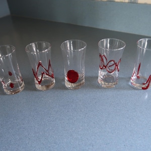 May include: Five clear shot glasses with red decorative accents. Each glass has a different design, including a red dot, swirls, and lines. The glasses are arranged on a blue surface.