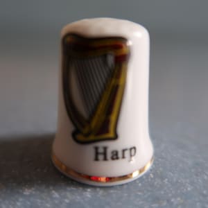 May include: A white ceramic thimble featuring a gold and brown harp design. The word "Harp" is printed in black at the base, with a gold trim around the bottom edge. A collectible item.