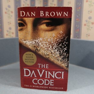 May include: A red paperback book with the title "The Da Vinci Code" by Dan Brown. The cover features a close-up of a painting with a person's eye and a piece of parchment with writing on it. The book is labeled as "Now a major motion picture" and "The #1 Worldwide Bestseller."