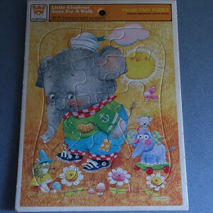 May include: A frame-tray puzzle featuring a cartoon illustration of a little elephant wearing a sailor hat and walking with a peanut in its trunk. The puzzle is set against a yellow background with a blue sky, a sun, and flowers. The puzzle is titled "Little Elephant Goes For A Walk".