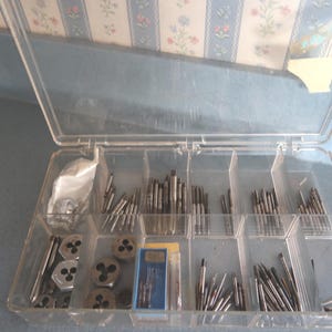 May include: A clear plastic toolbox filled with various metal tap and die tools. The tools are arranged in separate compartments, with a blue and white floral patterned wallpaper in the background.
