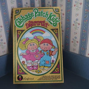 Peut inclure: Une boîte rectangulaire de Cabbage Patch Kids Colorforms Dress Up Set avec un fond jaune et une illustration ovale centrale de deux personnages, un arc-en-ciel et des fleurs. Le texte indique Cabbage Patch Kids Colorforms Dress Up Set.