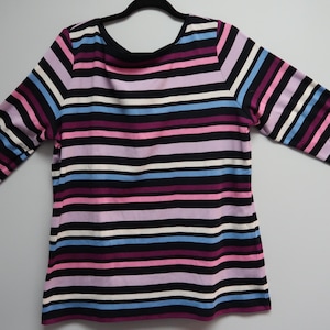May include: A long-sleeved striped shirt in a variety of colors, including black, white, pink, purple, and blue. The shirt is hanging on a black hanger against a plain white background. The sleeves are three-quarter length.