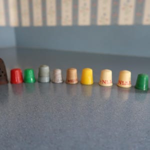 May include: A collection of colorful thimbles in various sizes. The thimbles are made of different materials and colors, including brown, red, green, gray, tan, yellow, and pink. Some thimbles have the letter "N" printed on them.