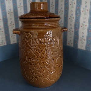 May include: A brown ceramic Rumtopf jar with a lid, handles, and embossed fruit designs. The jar features the word "Rumtopf" in script. The jar is cylindrical and sits on a blue surface.