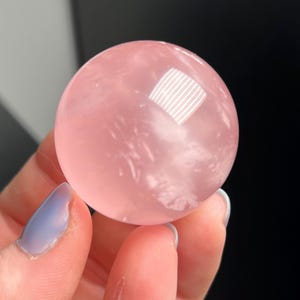 Peut inclure: Une sphère en cristal de quartz rose translucide. La boule polie est d'un rose tendre avec de subtiles veines internes. Le cristal reflète la lumière, créant un aspect clair et lumineux. Le fond est sombre, mettant en valeur la couleur et la forme du cristal.