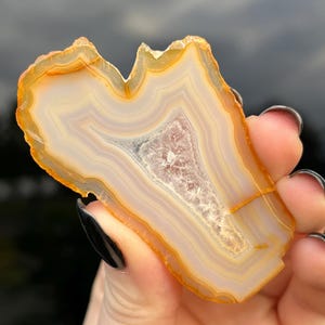 May include: A polished agate slice, displaying concentric bands of orange, white, and cream hues. The stone features a central cavity with crystal formations. The agate is held up against a cloudy sky, highlighting its natural beauty.