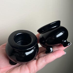 May include: Two glossy black miniature cauldron-shaped objects. Each cauldron has a rounded body, a wide opening, and three small feet. One cauldron is upright, while the other is tilted. The objects are displayed against a neutral background.