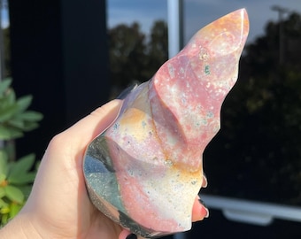 May include: A polished, freeform ocean jasper stone. The stone features a blend of colors, including pink, white, yellow, and black, with a swirling pattern. The stone is held in a hand, showcasing its unique shape and natural beauty.