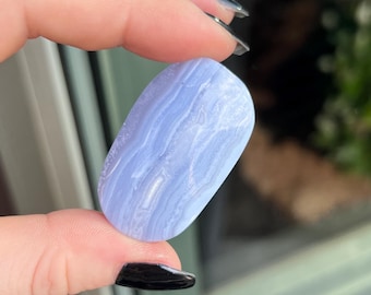 May include: A polished, oval-shaped blue lace agate gemstone. The stone has a light blue color with white, wavy banding patterns. The gemstone is held between fingers, showcasing its smooth, rounded form and natural beauty. This stone is a type of crystal.