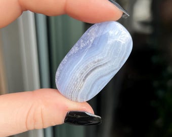 May include: A polished, oval-shaped blue lace agate stone. The stone features bands of light blue, white, and gray, creating a layered effect. The smooth surface reflects light, highlighting the natural patterns. The stone is held between two fingers.