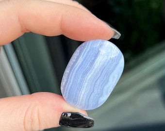 May include: A polished blue lace agate gemstone, oval-shaped with banded patterns. The stone is held between two fingers, showcasing its light blue and white layers. The gemstone is a natural crystal, often used in jewelry and healing practices.