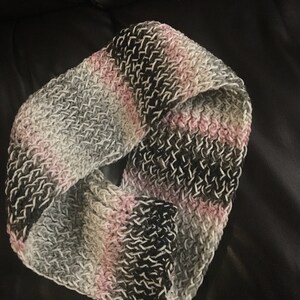May include: A crocheted infinity scarf in shades of gray, pink, and black. The scarf is made with a textured stitch and has a soft, cozy feel.
