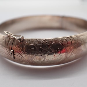 Pode incluir: Uma pulseira rígida articulada de tom prateado com um intrincado padrão floral e de volutas gravado na sua superfície. A peça circular inclui um mecanismo de fecho metálico seguro na lateral, permitindo abrir e fechar a pulseira facilmente.