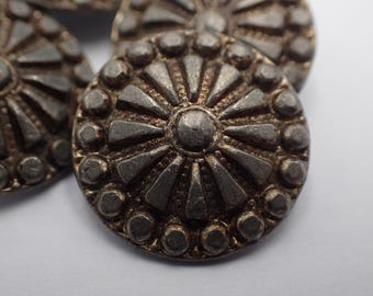 Op de afbeelding: Een close-up van een zilveren metalen knoop met een cirkelvormig ontwerp. De knoop heeft een verhoogd midden met een stervormig patroon en een ring van kleinere cirkels rond de rand.