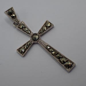 Könnte beinhalten: Silberfarbener Kreuzanhänger mit Markasitsteinen. Das Kreuz hat ein klassisches Design mit einem zentralen Stein und kleineren Steinen entlang der Arme. Der Anhänger ist wahrscheinlich aus Sterlingsilber und eignet sich für religiöse oder modische Zwecke.