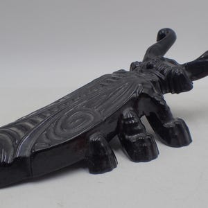 Może przedstawiać: Czarna, stylizowana rzeźba owada ze szczegółowymi skrzydłami i zakrzywionymi czułkami. Rzeźba ma sześć nóg i teksturowany korpus, przypominający cykadę. Dzieło sztuki jest prawdopodobnie wykonane z litego materiału.