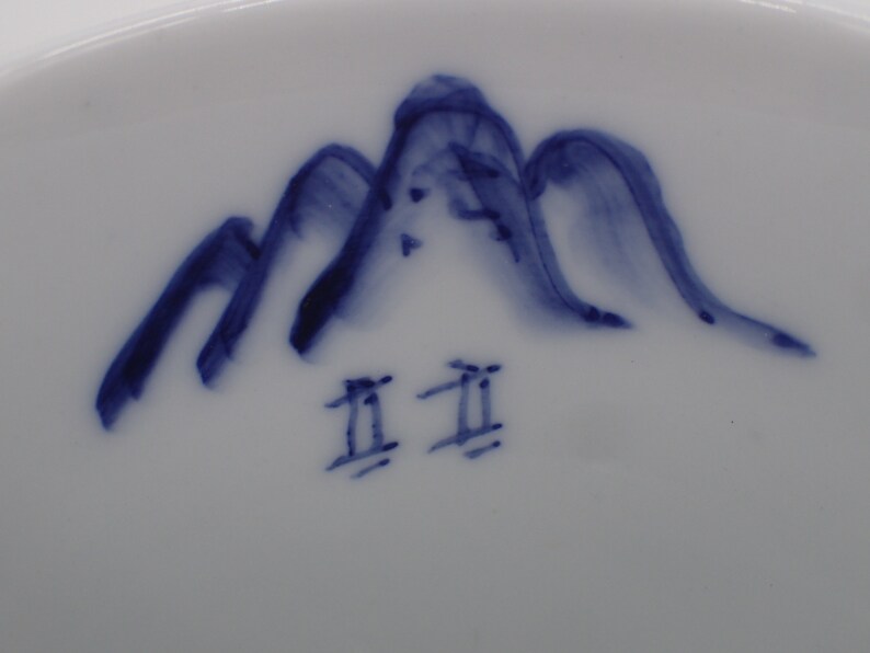 以下が含まれることがあります： 白地に青色の山と「工  工」の文字が描かれた白い陶器の皿のアップ写真です。