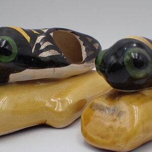 Peut inclure: Deux figurines de grenouilles en céramique. Les grenouilles ont des têtes noires avec des yeux verts et des détails à motifs jaunes et blancs. Elles reposent sur des bases oblongues jaunes. Ce sont probablement des objets décoratifs.