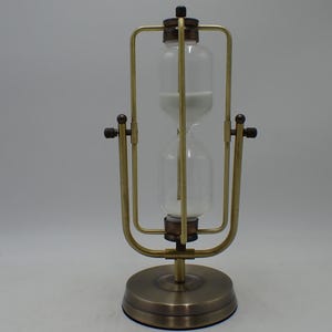 Pode incluir: Uma ampulheta rotativa com um recipiente de vidro transparente preenchido com areia branca, inserida numa estrutura metálica de tom latão com uma base circular. A estrutura inclui um mecanismo de pivô central para virar a ampulheta.