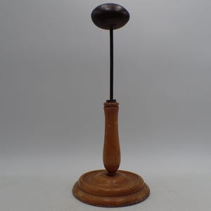 May include: A vintage hat stand with a wooden base and stem, topped with a dark brown, oval-shaped head. The stand has a classic design, perfect for displaying hats. The base is approximately 15 cm in diameter.