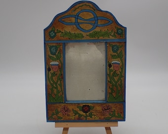 May include: A wooden picture frame with a blue and gold painted design. The frame features a Celtic knot design at the top and floral motifs on the sides. The frame is standing on a small wooden easel.