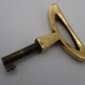 May include: A brass key with a unique, abstract design. The key has a long, thin shaft and a large, decorative head. The head is shaped like a stylized letter T with a curved top.