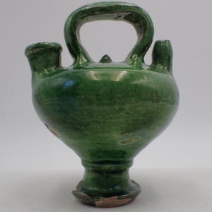 May include: A green ceramic jug with a large handle, two spouts, and a rounded body. The jug has a glossy finish and a pedestal base. The overall design is reminiscent of antique pottery.