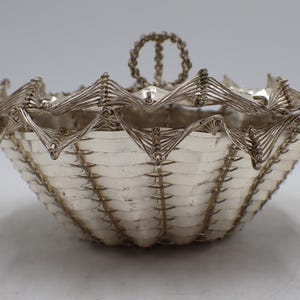 Può includere: Un cestino color argento con un design intrecciato e un bordo smerlato decorativo. Il cestino ha un manico nella parte superiore ed è realizzato in metallo. Il design del cestino è intricato e dettagliato, con una superficie strutturata.