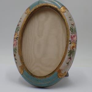 May include: An oval picture frame featuring a light blue and cream-colored border with hand-painted floral motifs in pink, yellow, and green. The inner edge is lined with a textured gold-tone metallic trim, surrounding a plain cream-colored insert.