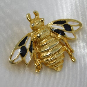 Puede incluir: Broche de abeja dorada con alas de esmalte negro y blanco. La abeja tiene un cuerpo detallado y texturizado.