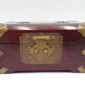 May include: A rectangular wooden box with a dark reddish-brown finish. The box features ornate brass corner pieces, a central latch, and decorative accents. The brass hardware adds a vintage aesthetic to the box.