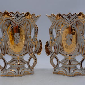 Pode incluir: Um par de vasos de cerâmica branca com designs intrincados de filigrana, bordas recortadas e detalhes em ouro metálico. Cada vaso exibe um painel oval central com um motivo floral branco sobre um fundo dourado reflexivo.