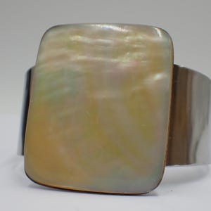 Könnte beinhalten: Breites silbernes Armband mit einem großen, rechteckigen, schillernden Perlmutt-Mittelstück. Das Perlmutt hat einen perlmuttartigen Glanz mit Gelb-, Grün- und Weißtönen. Das Armband ist ein schlichtes, elegantes Design.