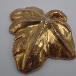 May include: A gold-colored leaf-shaped decorative object. The leaf has a textured surface with visible veins and a metallic sheen. The object is likely made of metal or a similar material and is designed for decorative purposes.