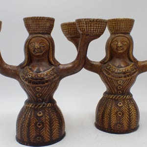 May include: A pair of brown ceramic candle holders shaped like figures with outstretched arms. Each figure wears a textured dress and supports a basket-like candle cup on their head, with additional candle cups extending from their hands.