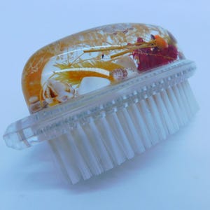 Könnte beinhalten: Eine transparente Plastik-Nagelbürste mit weißen Borsten. Der Griff ist mit einem durchsichtigen Harz-Oval verziert, das getrocknete Blumen und Muscheln enthält.