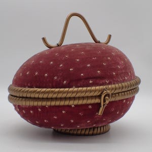 以下が含まれることがあります： ブルゴーニュ色の卵型の小物入れ。金色のハンドルとトリムが付いています。ボックスは、小さな白い水玉模様のベルベットのような素材で覆われています。トリムは、ねじれたロープのデザインです。