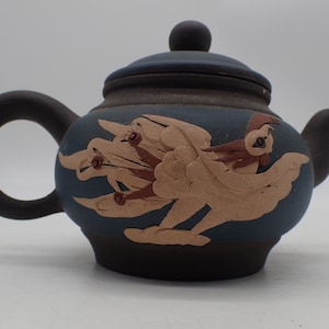 May include: A small, dark blue teapot with a brown lid and a raised design of a bird with brown accents.
