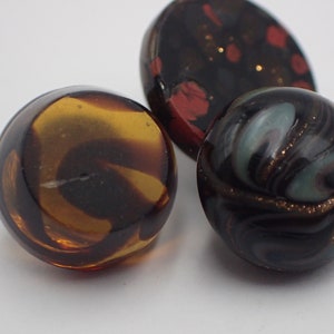 Op de afbeelding: Drie ronde glazen knopen met verschillende patronen. Eén knoop is bruin en amberkleurig met swirls, één is zwart met rode en gouden vlekken en één is zwart met draaiende witte en gouden accenten.
