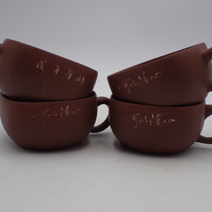 May include: Set of four brown ceramic teacups with handles. Each cup has Chinese characters inscribed on the side.