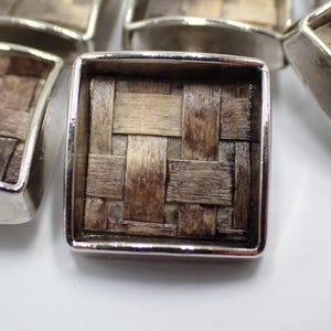 May include: Close-up of several square silver-toned metal buttons. Each button features a woven, basket-like design in shades of brown and beige. The buttons are arranged on a white surface, with some slightly out of focus.