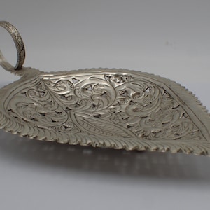 May include: A silver serving dish shaped like a leaf with intricate floral designs and a ring handle.