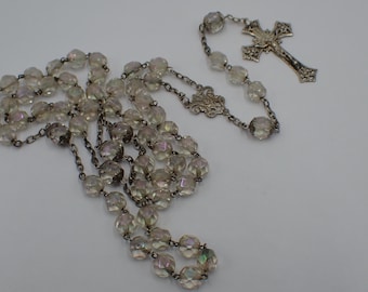 Könnte beinhalten: Ein Rosenkranz mit irisierenden, facettierten Perlen und einem silberfarbenen Kreuzanhänger. Die Perlen sind auf einer filigranen Kette aufgereiht, und das Kreuz weist aufwendige Details auf. Ein religiöses Objekt.