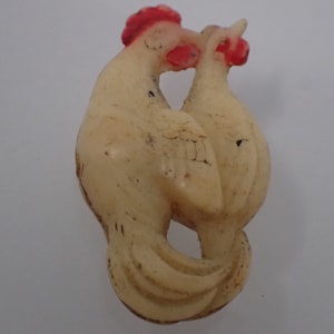 Könnte beinhalten: Eine weiße Keramikfigur von zwei Hähnen, einer mit dem Kopf zur Seite gedreht und der andere mit dem Kopf nach vorne gedreht. Beide Hähne haben rote Kämme und Kehllappen.