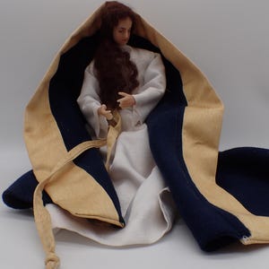 Peut inclure: Une poupée vêtue d'une robe blanche et d'une cape à capuche beige et bleu marine. La poupée a de longs cheveux roux ondulés. La cape a un extérieur beige et un intérieur bleu marine avec une longue cravate beige. La poupée est posée en position assise.