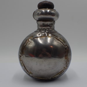 May include: A vintage silver-colored flask with a rounded body and a decorative design. The flask has a small neck and a dark brown stopper. The metal shows signs of age and wear, adding to its antique charm.